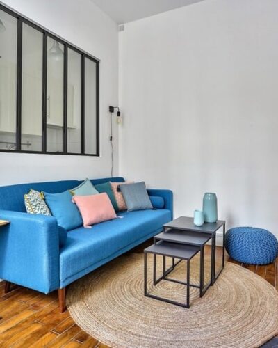 Salon moderne avec canapé bleu, verrière donnant sur la cuisine, cheminée en marbre et parquet d'origine dans un deux-pièces situé dans le quartier Blanche, Paris 9e.