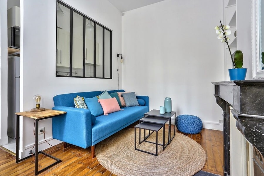 Salon moderne avec canapé bleu, verrière donnant sur la cuisine, cheminée en marbre et parquet d'origine dans un deux-pièces situé dans le quartier Blanche, Paris 9e.