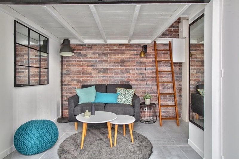 Salon avec murs en briques apparentes, canapé moderne, échelle menant à une mezzanine, situé dans un studio à Montmartre, Paris 18e, proposé par un chasseur immobilier expert à Paris.