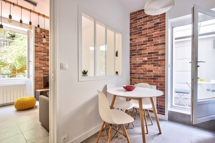 Coin repas avec table ronde et verrière, murs en briques apparentes et accès à une terrasse, situé dans un appartement à Asnières, proche de la gare, proposé par un chasseur immobilier dans les Hauts-de-Seine.