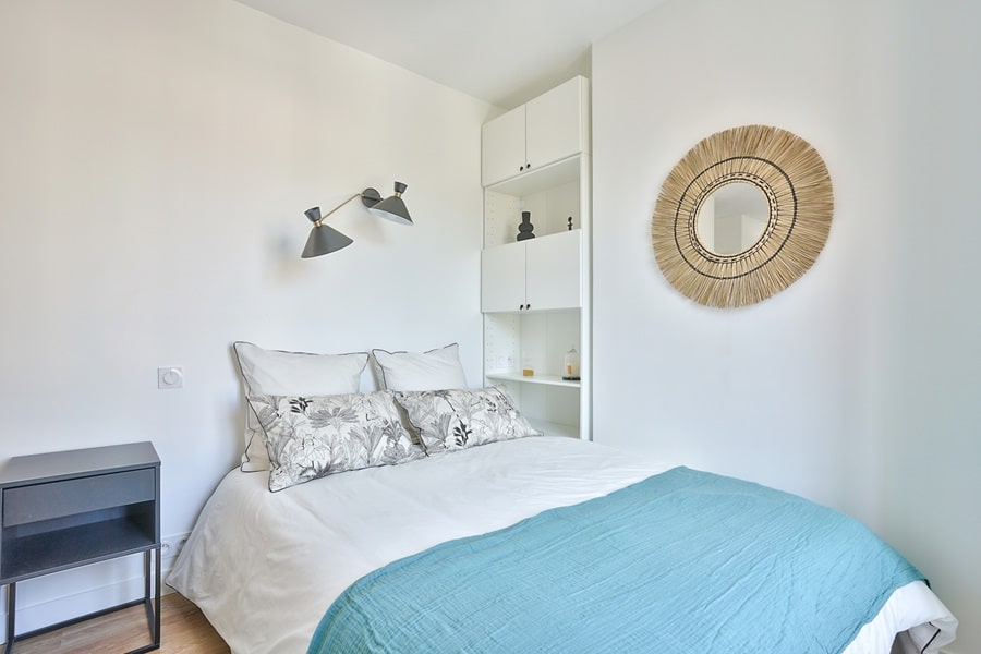 Chambre d’un studio clé en main à Asnières-sur-Seine avec une décoration minimaliste, une applique murale noire, un miroir en rotin et une literie élégante.