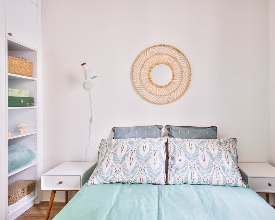 Chambre lumineuse avec une décoration raffinée, incluant un miroir en rotin, des coussins à motifs modernes et des rangements intégrés dans un appartement parisien.