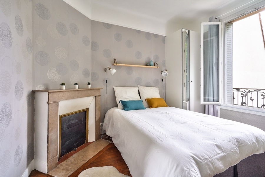 Chambre décorée avec un papier peint à motifs, une cheminée d’époque, une étagère en bois flotté, et un lit confortable avec vue sur un balcon, située dans un appartement deux-pièces à La Garenne-Colombes.