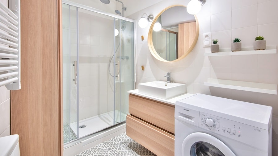 Salle de bain lumineuse avec une douche italienne, meuble en bois clair, miroir rond, et lave-linge intégré dans un appartement du 9e arrondissement de Paris.