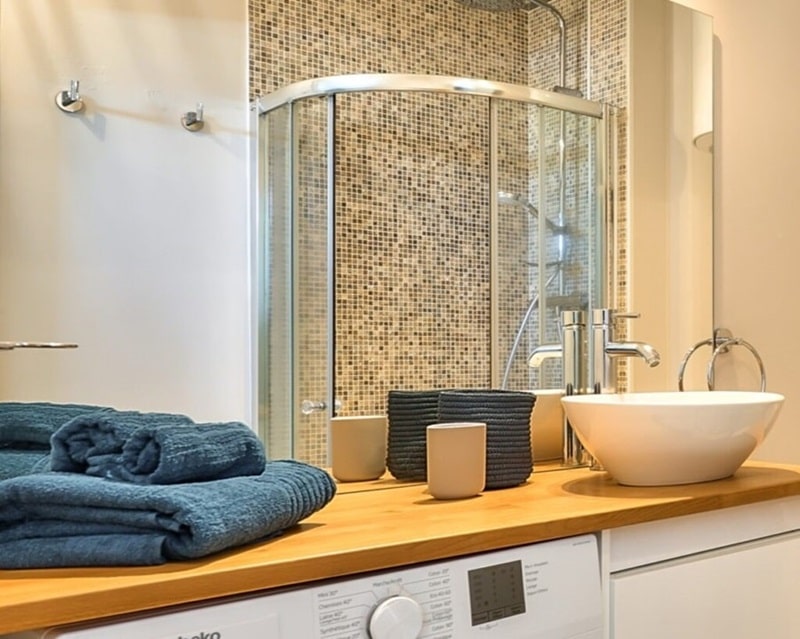 Salle de bain avec douche en mosaïque, vasque design et plan de travail en bois, équipée d’une machine à laver, sélectionnée par Early Birds, chasseurs immobiliers spécialisés à Paris et ses environs, Rue Sébastopol à La Garenne-Colombes.