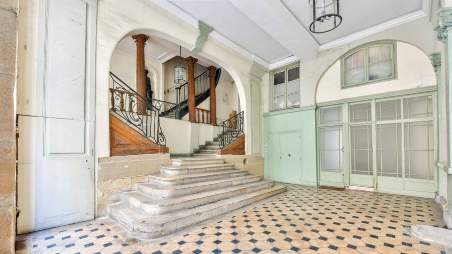 Hall d'entrée spacieux d'un immeuble parisien avec des escaliers en pierre, des colonnes sculptées, des carreaux en mosaïque et des portes vitrées de style ancien.