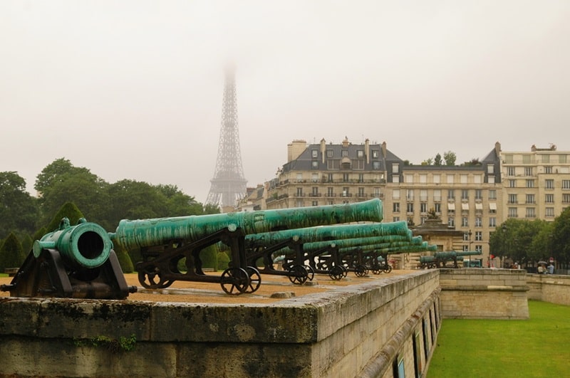Les canons exposés aux Invalides à Paris, avec une vue brumeuse sur la Tour Eiffel en arrière-plan.